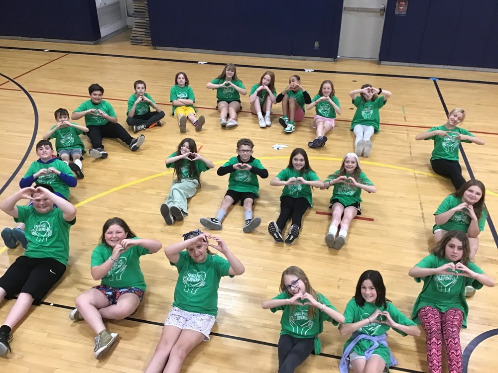 A large group of students in green t-shirts sitting on the gym floor, making the caring sign with their hands